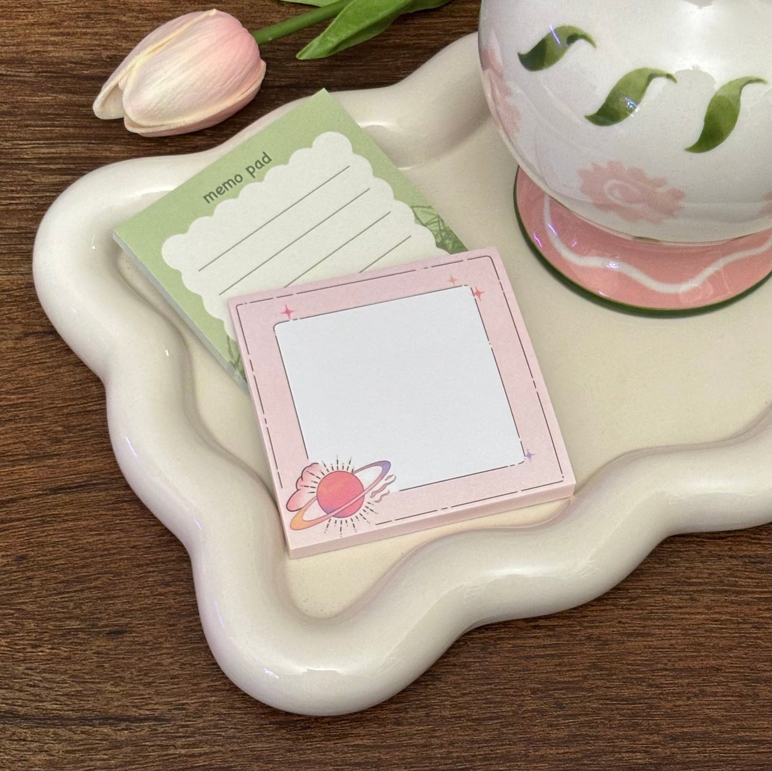 Cute pastel memo pads on a wavy cream tray beside a floral ceramic mug and pink tulip, featuring a green lined notepad and a pink square sticky note with a planet illustration.