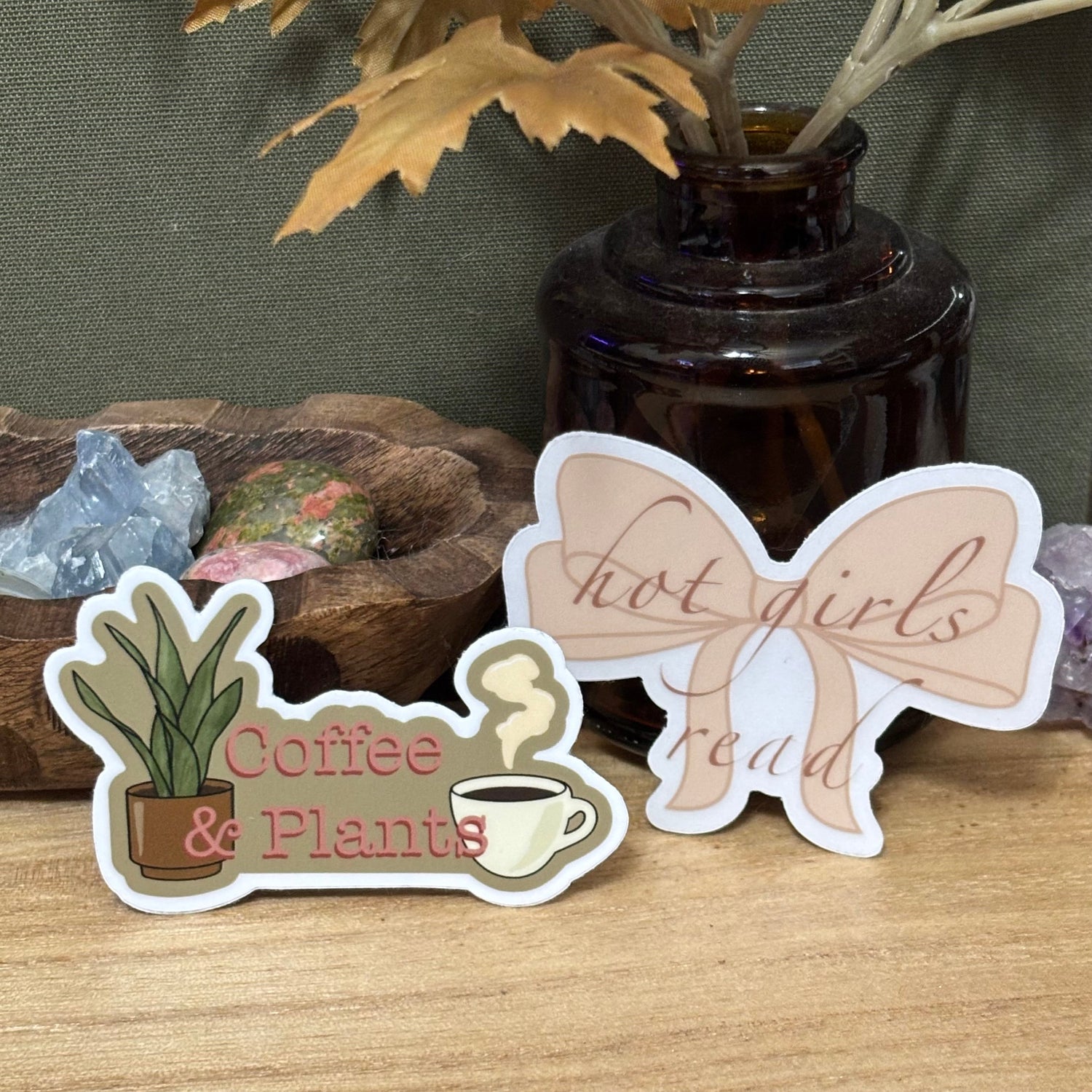 Aesthetic coffee and plant themed stickers including “Coffee & Plants” with a potted plant and mug and a bow shaped “hot girls read” sticker, styled on a wooden desk with crystals and dried leaves.